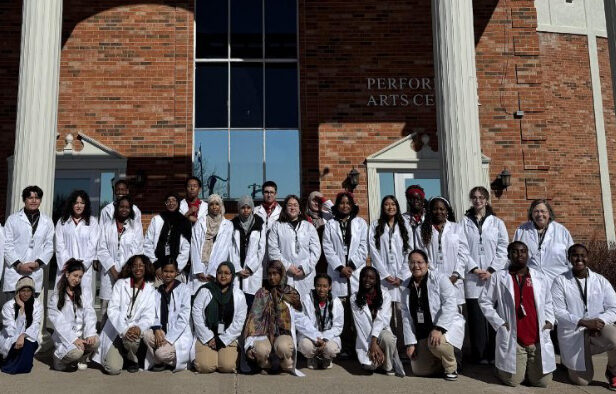 A group of many students in white lab coats outside a brick building.